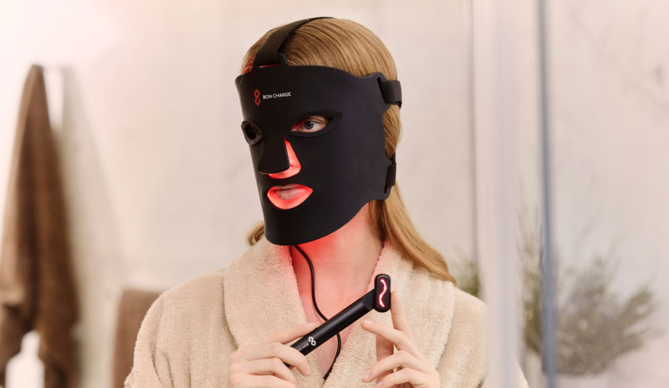 Woman in bathroom wearing a Red Light Face Mask, while holding a Red Light Face Wand